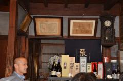 棚に様々なお酒が置かれてあり、その上に表彰状と振り子時計が置かれてある老舗・張替酒屋の内部の写真