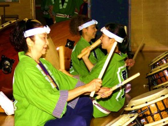 緑色の法被と頭に手ぬぐいハチマキを巻いた3名の女性がバチを持って和太鼓を叩いている写真