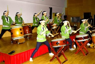 緑色の法被を着て頭に手ぬぐいハチマキを巻いた数名の女性がステージで和太鼓を披露している様子の写真