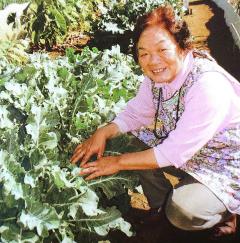 畑に植えられている野菜の横に座っている桜井勝子さんの写真