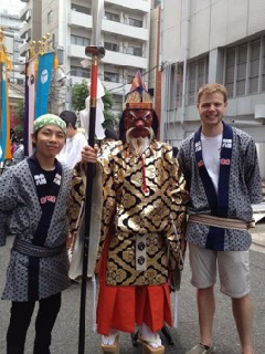 山伏の衣装を着て天狗のお面を付けた人の左横に法被を着たブラックウッドさん、右横に法被姿の人と3人で記念撮影を行っている写真