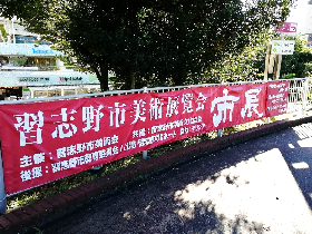 赤い横断幕に白字で「習志野市美術展覧会 市展」と書かれた横断幕が、道路沿いのフェンスに張られている写真