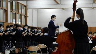 津田沼高等学校オーケストラ部が演奏をし、城東高校の生徒たちが楽譜を持ち歌を歌っている様子の写真