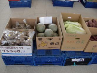 青いかごの上に並べられたダンボールの箱に入ったサツマイモや白菜などの野菜の写真