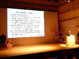 ステージ上の文章が映し出されたスクリーンの横に立ち話をしている様子の山岸先生の写真
