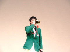 緑色のジャケットを羽織り、右手でラッパを持ち演奏しているトリトン海野さんの写真