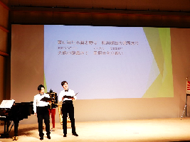 文字が映し出されたプロジェクタースクリーンのあるステージ上で木村さんと喜久田さんが譜面を持ち歌っている写真
