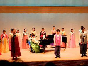 韓国の民族衣装を着た人たちがピアノを囲んで歌を歌っている舞台の上の写真