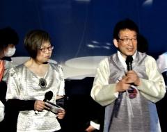 シルバーの衣装を着た実行委員会戸田さんと指揮者の山岡先生がマイクで挨拶をしている写真