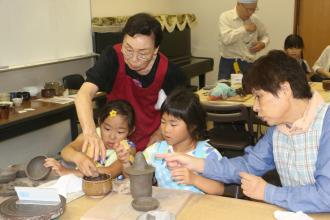 粘土をこねて器を作っている2人の子供と傍でサポートしている2名の女性の写真