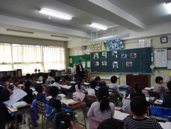 前方の黒板に資料を貼り、授業を行っている先生と子供たちを後方から写した写真