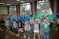 子供達が置くからオレンジ、水色、青、緑のボンボンを両手に持ち、両手を上にあげてダンスを披露している写真