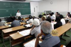 黒板の前に立ち話をしている先生と席について講義を聞いている参加者の写真