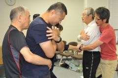 男性を後ろから抱きかかえるような形で腹部突き上げ法の訓練を行う参加者の方々の写真