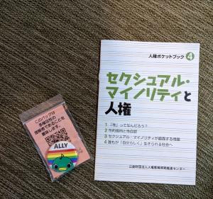 「セクシャルマイノリティと人権」と書かれた冊子と、キャラクターが描かれた虹色の缶バッジのような物が置かれている写真