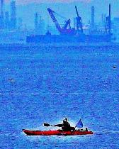 遠くに大きな船舶や工場地帯が見える海上を漕ぎ進むカヌーの写真