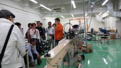 千葉工業大学の室内で大きな機械の前で説明をするオレンジ色の服を着た男性と説明を聞いている受講者の方々の写真