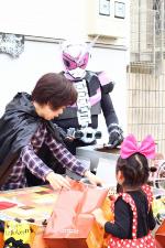 仮面ライダーに仮装した大人の人とローブを着た女性にお菓子を貰っている子供の後ろ姿の写真
