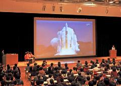 舞台上の大きなスクリーンに発射するロケットの映像が映し出されている写真