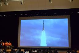 白煙を噴射しながら垂直に飛んでいくロケットの映像が映し出されたスクリーンの写真