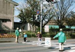 小学校の近くの横断歩道で2名の交通安全ボランティアが小学生の登校を見守っている写真