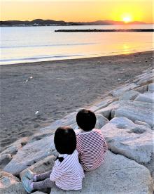 海岸の岩の上で子ども2人が水平線上の太陽を眺めている後ろ姿の写真