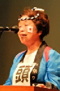 法被を着て頬に日本国旗のマークを付けた林崎実行委員長が壇上でスピーチしている写真