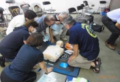 AEDの機械と訓練用のマネキンの周りに集まって講習を受けている参加者と指導員の写真