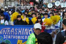 青い横断幕を先頭に町中を行進しているチアリーダーと習志野高校吹奏楽部の写真