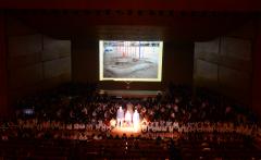 舞台上のスクリーンに写真が映し出されて参加者と共に震災を振り返っている式典の様子の写真