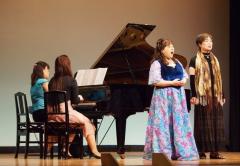 グランドピアノを弾いている2名の女性と演奏する音楽に合わせてドレスを着た2名の女性が歌を歌っている写真
