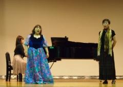 演奏するピアノの前でドレスを着た女性2名が歌を披露している写真