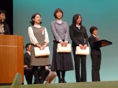 ステージ上で3名の標語コンクールの受賞者が賞状を手に持ち並んで立っている写真