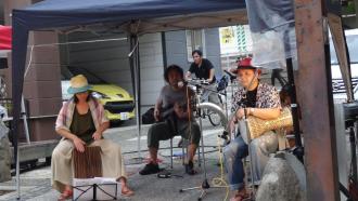 テントの下で3名のアーティストが椅子に座り打楽器などの楽器を演奏している駅前でのライブの様子の写真