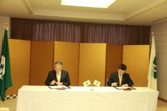スーツ姿の2名の男性が、金屏風の前で京田辺市と習志野市の協定書に押印している写真