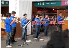 青色のシャツを着た人達がロビーで管楽器の演奏を行っている様子の写真