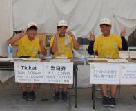 お揃いの黄色いTシャツに帽子を被った、チケット売り場担当のボランティアの方3名が笑顔でピースサインをしている写真