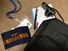 腕章、カメラ、名札、ノート、2本のペンが置かれている取材道具の写真