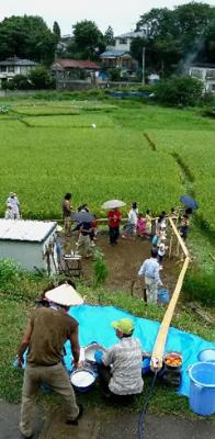 緑色の田んぼの上から孟宗竹で造った流しそうめん台と、そこからそうめんを流そうと待機している男性と、下で待つ人達を上から写した写真