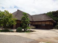 晴天時に撮影された茅葺の旧鴇田家住宅と木の植えられた庭の写真