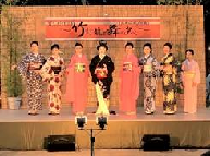 色々な柄の着物を身に着けた8名の女性が舞台上に並んでいる写真