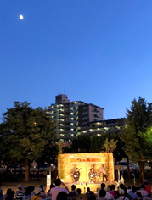 空にはオレンジ色の月が斜め左上空にあり、後方には構想ビル、手前には津軽三味線を演奏している舞台と来場者の方々が写っている祭りの風景写真