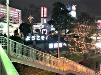 歩道橋から見える「ようこそ！津田沼へ」の大きな文字照明とイルミネーションで輝く街並みの様子の写真