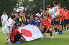 応援メッセージを描いた日本国旗を囲み、小学生と代表選手が記念撮影をしている所を斜め前から写した写真