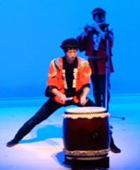 長胴太鼓の前でオレンジ色の法被を男性がバチを持って、足を大きく広げ腰を下ろした姿勢で太鼓の演奏を披露している写真