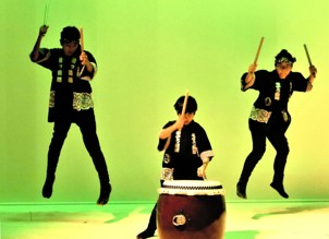 中央で長胴太鼓を叩いている演者と後方の2人の演者がバチを持って飛び上がった瞬間を写した演奏の様子の写真