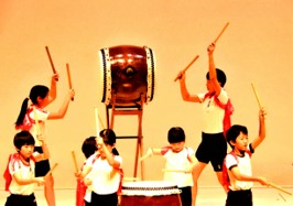 舞台上で台座の上の和太鼓を向い合って叩いている児童、長胴太鼓をバチを振り上げて叩いている児童達の和太鼓演奏の様子の写真