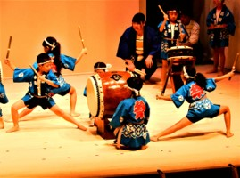 青い法被を着た児童達がバチを両手に持ち、腰を下ろして左脚を前に出して構え、和太鼓の演奏を披露している写真