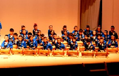 青い法被を着た児童達が竹太鼓の前に並んで座り、演奏を披露している写真