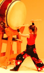 台座の上に乗せられた大きな平太鼓を女性の演奏者がバチを振り上げて力強い演奏を披露している写真